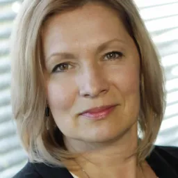 Portret uśmiechniętej kobiety o blond włosach, w czarnej marynarce i białej bluzce, na tle okna z żaluzjami.