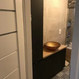 Nowoczesna łazienka z czarną szafką pod umywalkę, drewnianym blatem i okrągłą, miedzianą umywalką na tle beżowych płytek i mozaikowej podłogi.