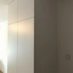 Wysoka, biała szafa wnękowa z czterema drzwiami, zlicowana ze ścianą, widoczna w narożniku pomieszczenia.