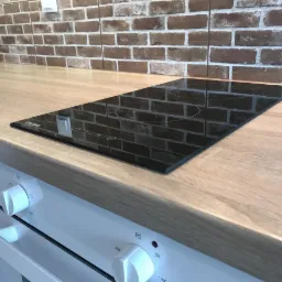 Płyta indukcyjna zamontowana w blacie w kuchni w Poznaniu, widoczny piekarnik z białymi pokrętłami i ceglana ściana w tle.