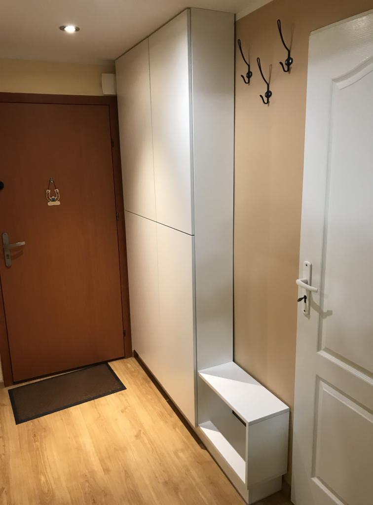 Wysoka, biała szafa z siedziskiem w przedpokoju, obok drzwi wejściowych i wewnętrznych, na ścianie wieszaki na ubrania.