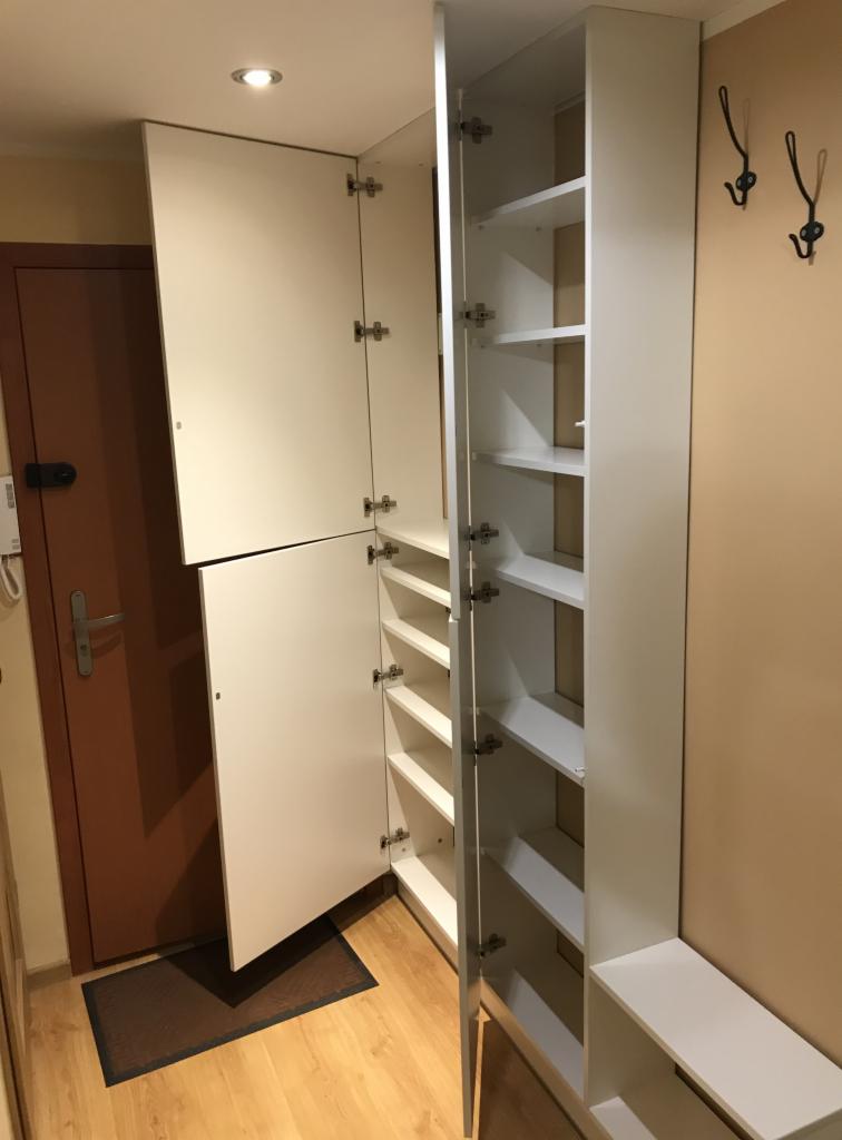 Biała szafa z półkami w przedpokoju, częściowo otwarta, widoczne zawiasy, obok brązowe drzwi wejściowe i wieszaki na ścianie.