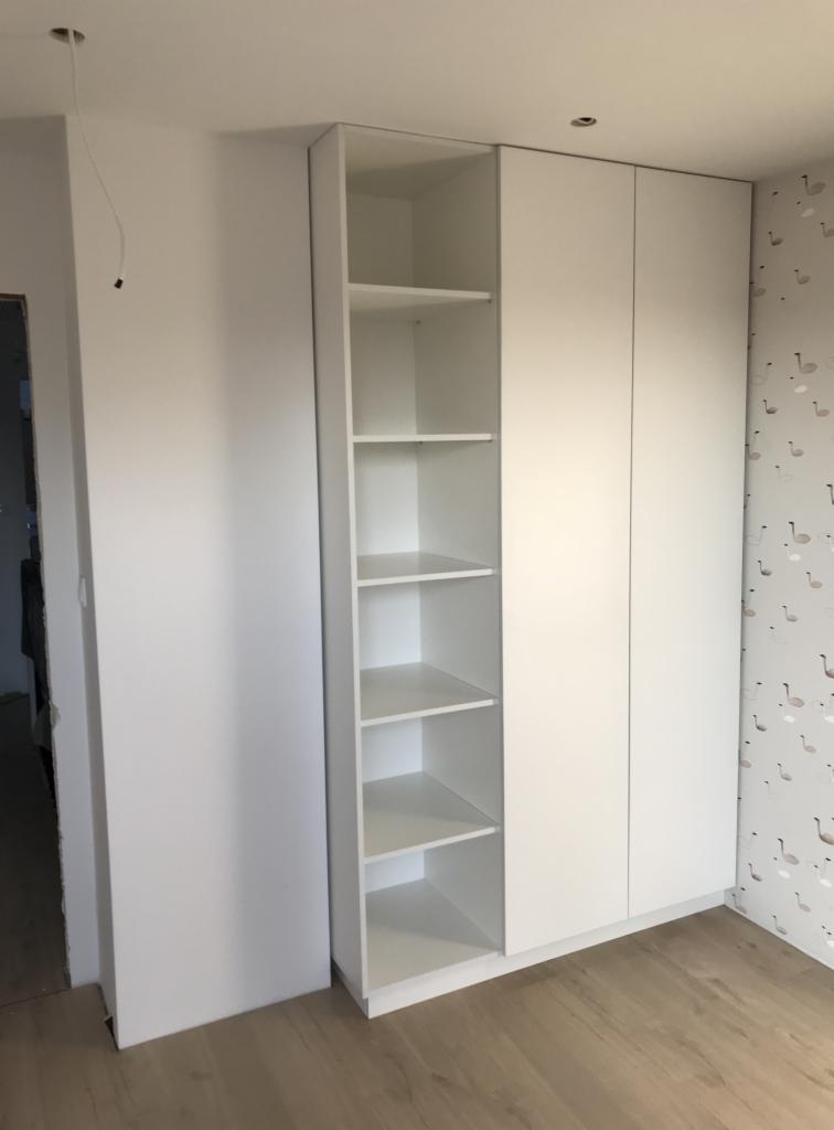 Biała szafa wnękowa z otwartą półką na tle ściany z tapetą w łabędzie, widoczny fragment podłogi z paneli.