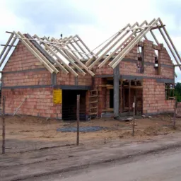 Surowy stan domu z czerwonej cegły z widoczną drewnianą konstrukcją dachu i otwartym garażem, ogrodzony tymczasowym płotem z drewnianych słupków.