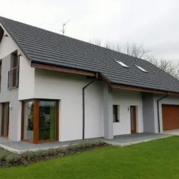 Nowoczesny dom jednorodzinny z grafitową dachówką ceramiczną, drewnianą stolarką okienną i garażem wbudowanym w bryłę budynku.