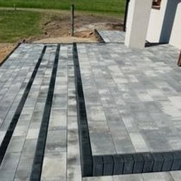 Usługi Ogólnobudowalne Andrzej Brygała - Nowoczesny taras z szarej kostki brukowej, z ciemnymi pasami i obrzeżami, prowadzący do wejścia domu z białymi kolumnami. Widok z góry, słoneczny dzień.