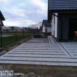 Ułożona nawierzchnia z szarej kostki brukowej w geometryczny wzór, widoczny fragment ogrodzenia z siatki i pracownik w garażu w tle.