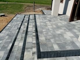 Nowoczesny taras z szarej kostki brukowej, z ciemnymi pasami i obrzeżami, prowadzący do wejścia domu z białymi kolumnami. Widok z góry, słoneczny dzień.