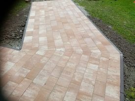 Ułożona, beżowa kostka brukowa w kształcie prostokątów tworząca ścieżkę w ogrodzie. Widoczne obrzeża i trawa. Kostka ułożona w jodełkę.