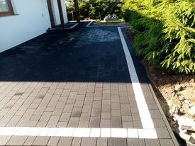 Nowoczesna nawierzchnia z kostki brukowej w odcieniach szarości, obramowana białym pasem, przylegająca do białego budynku z widocznym fragmentem ogrodu.