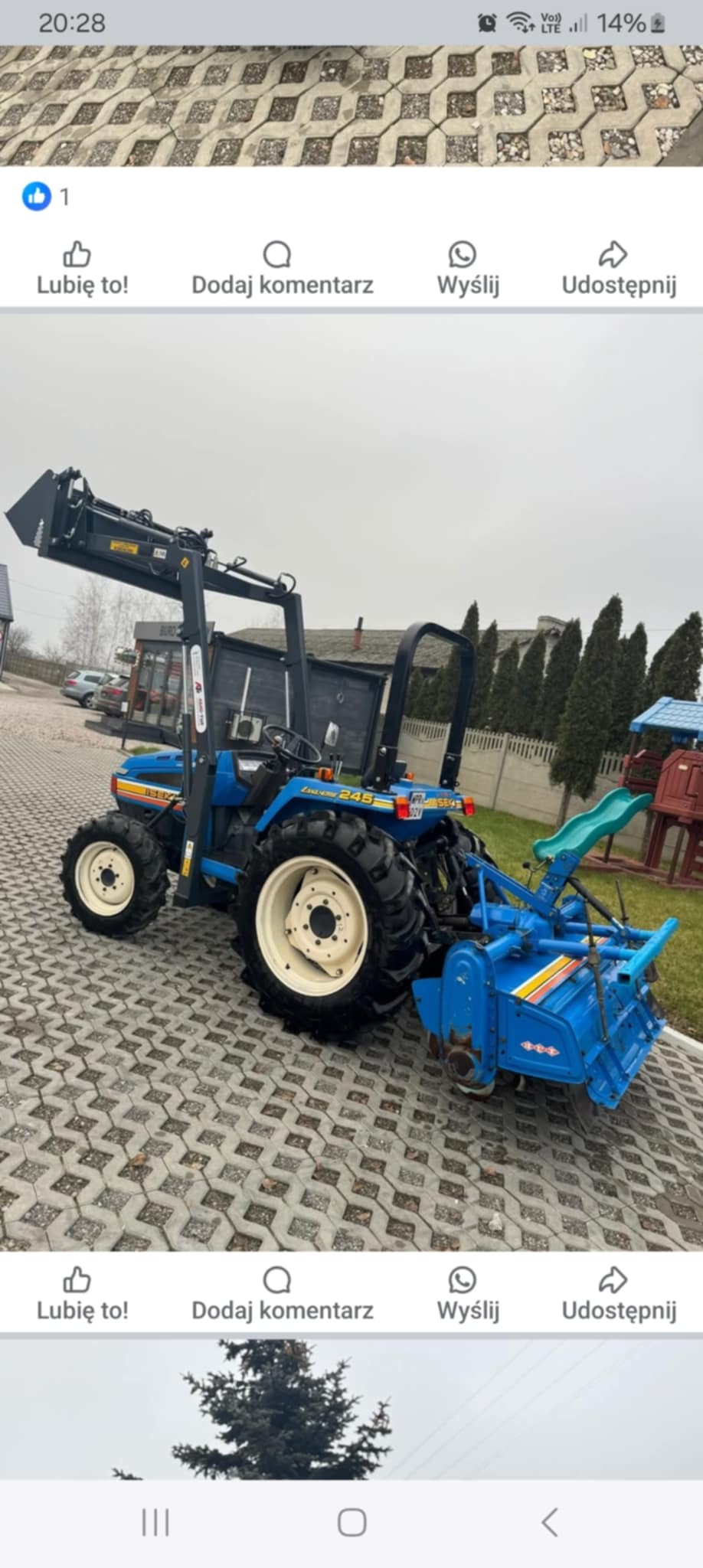 Niebieski traktor z glebogryzarką i ładowaczem czołowym na kostce brukowej. Widoczne budynki w tle. Sprzęt rolniczy gotowy do pracy.