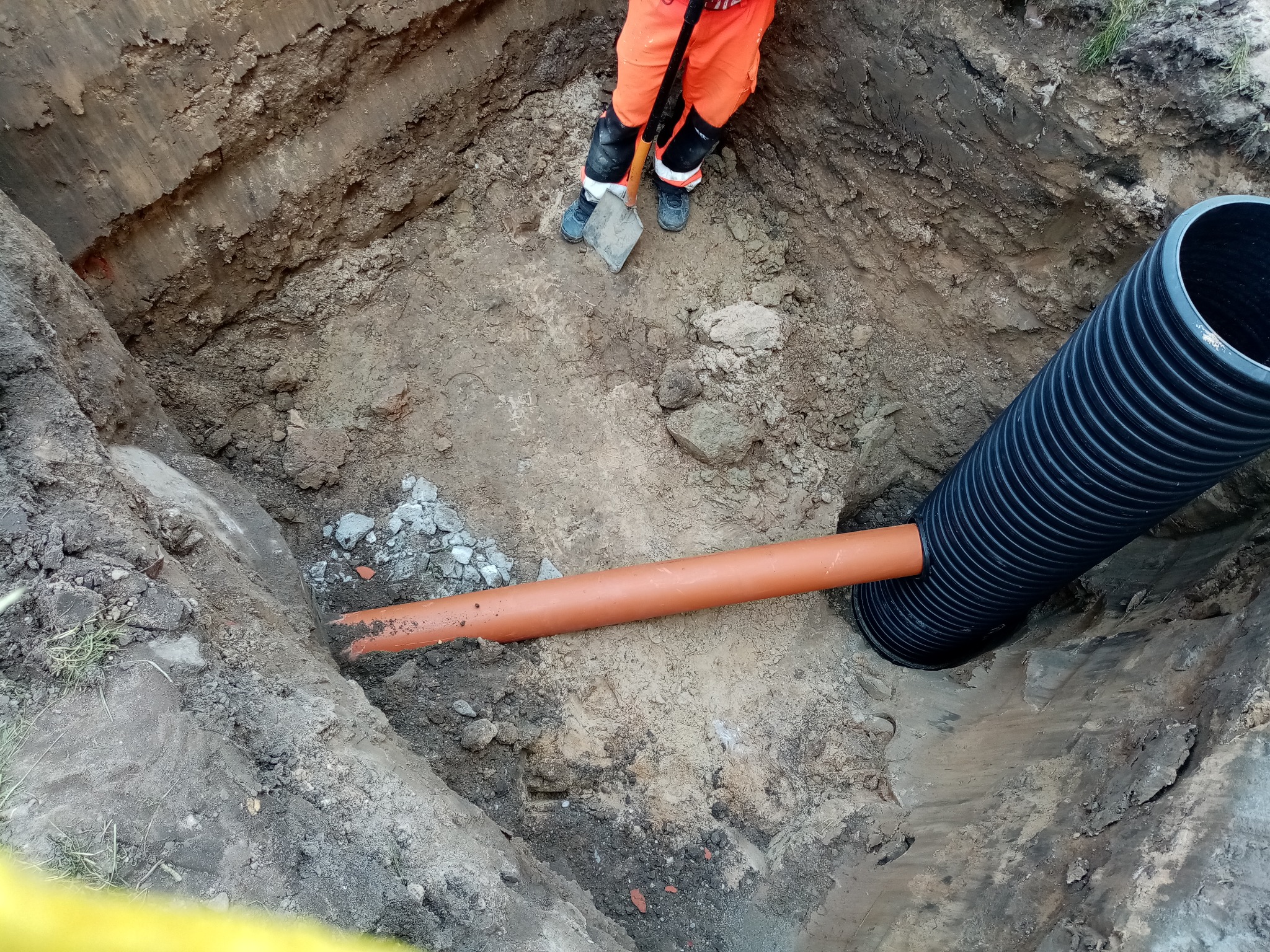 Wykop z ułożonymi rurami kanalizacyjnymi, widok z góry. Pracownik w pomarańczowym kombinezonie stoi w wykopie z łopatą. Rury czarne karbowane i pomarańczowe gładkie.