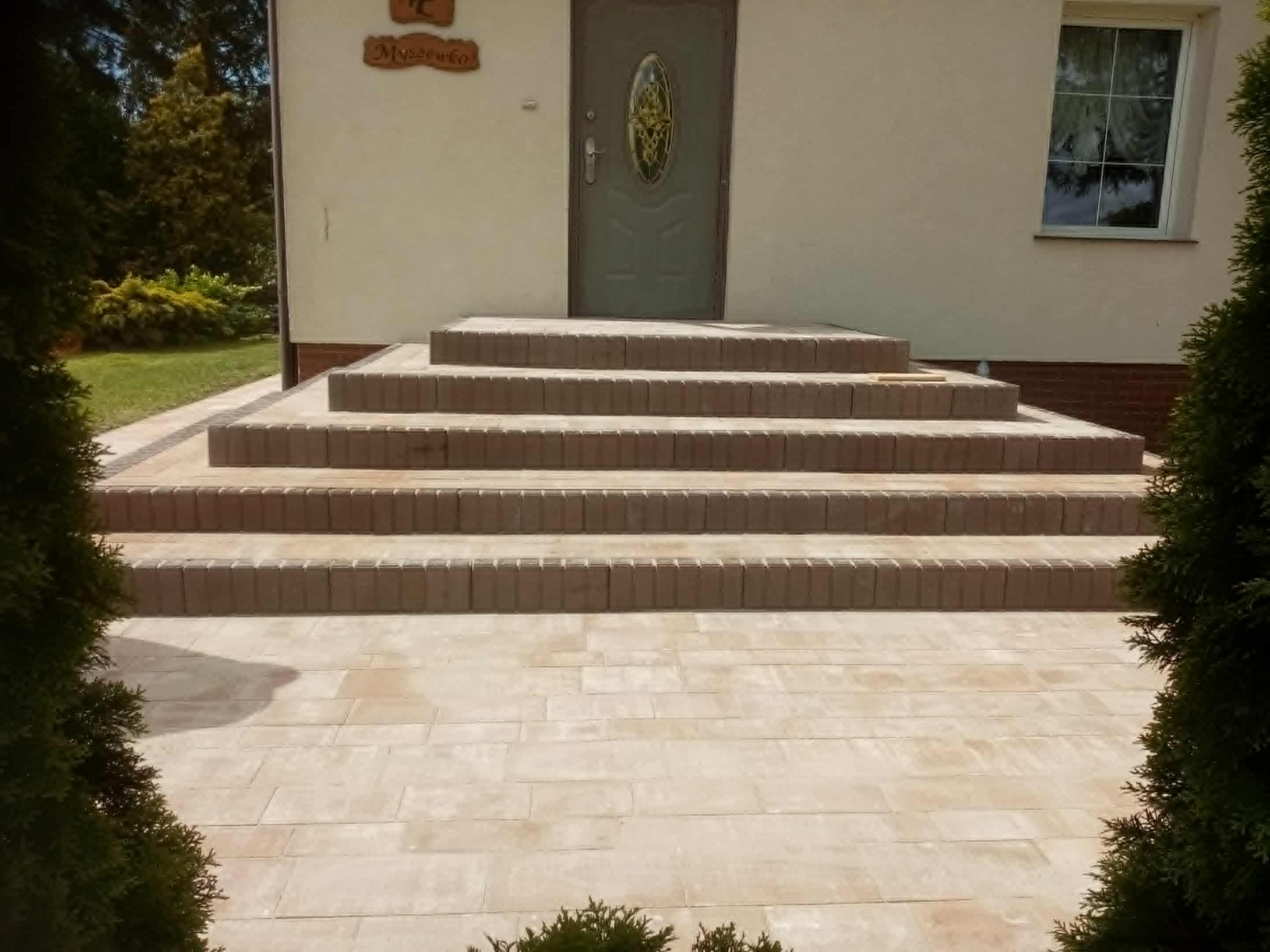 Jasne, prostokątne płyty chodnikowe prowadzą do czterech stopni z brązowej kostki, tworząc wejście do domu z szarozielonymi drzwiami i oknem po prawej stronie.