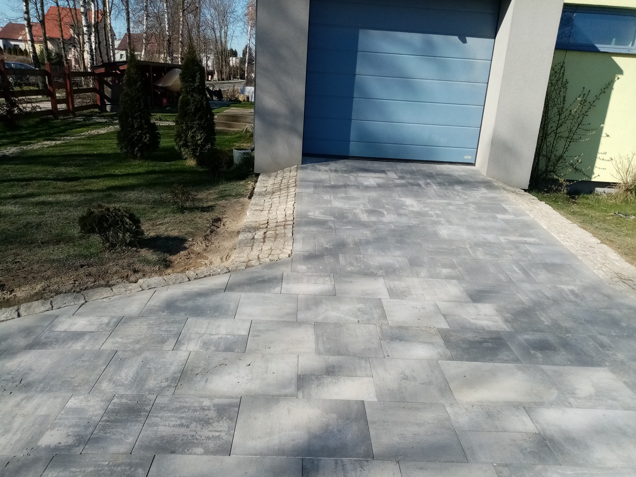 Szara kostka brukowa ułożona przed garażem z niebieską bramą, obrzeża z jasnego kamienia. Widoczne drzewa i krzewy w tle. Słoneczny dzień.