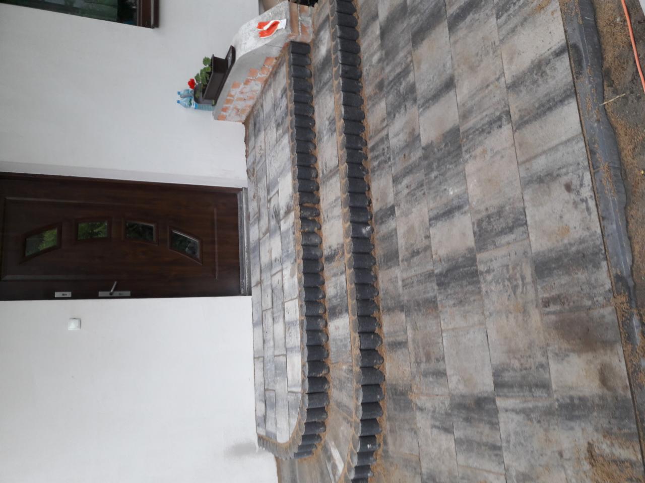 Wykończona elewacja budynku z szarej kostki brukowej z ciemnymi obrzeżami, widoczna część drzwi wejściowych i dekoracje na ścianie.