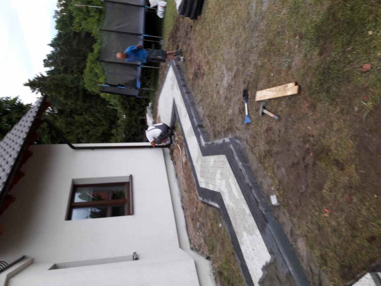 Pracownik układa zygzakowaty wzór z jasnoszarej kostki brukowej z ciemnoszarym obramowaniem wzdłuż ściany domu; w tle trawnik, trampolina i drugi pracownik.