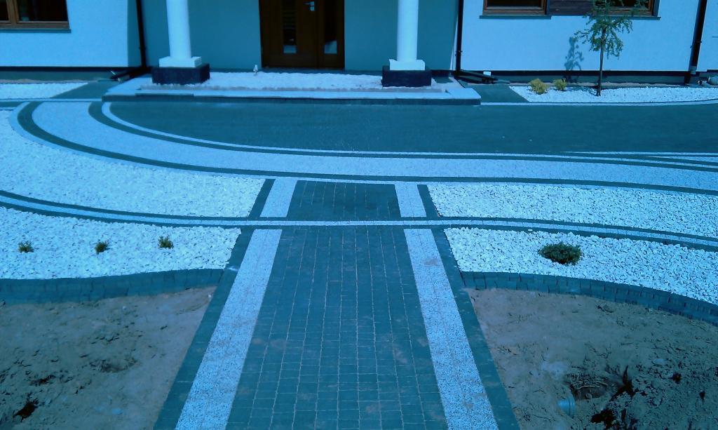 Nowoczesny podjazd z kostki brukowej w Nowym Dworze Gdańskim, z białymi kamieniami i elementami zieleni, prowadzący do wejścia domu z kolumnami.