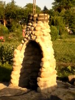 Murowany grill ogrodowy z kamienia polnego, z widocznym paleniskiem i kominem, otoczony zielenią ogrodu. Na dole widoczne niedociągnięcia w postaci resztek cementu.