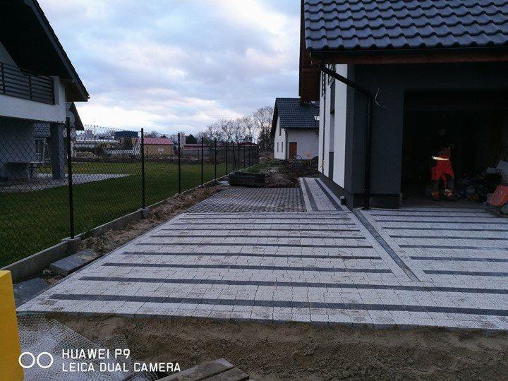 Ułożona nawierzchnia z szarej kostki brukowej w geometryczny wzór, widoczny fragment ogrodzenia z siatki i pracownik w garażu w tle.