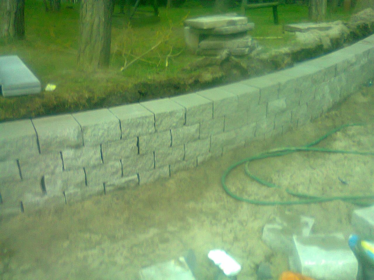 Niska, zakrzywiona ścianka oporowa zbudowana z szarych, betonowych bloczków, widoczna na tle trawy i drzew, z zielonym wężem ogrodowym leżącym obok.