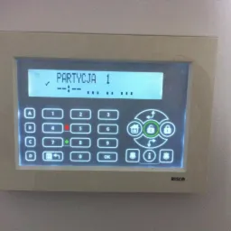 Panel sterowania alarmem firmy RISCO, wyświetlający komunikat 'PARTYCJA 1' i klawiaturę numeryczną z podświetlonymi przyciskami funkcyjnymi, zamontowany na jasnej ścianie.