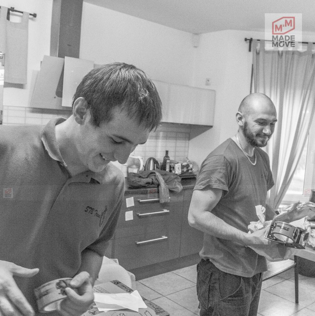 Dwóch mężczyzn uśmiechniętych podczas pakowania przedmiotów w kuchni, jeden owija taśmą pudełko, drugi trzyma zapakowany przedmiot, w tle widoczne szafki kuchenne i okno z zasłonami, zdjęcie...