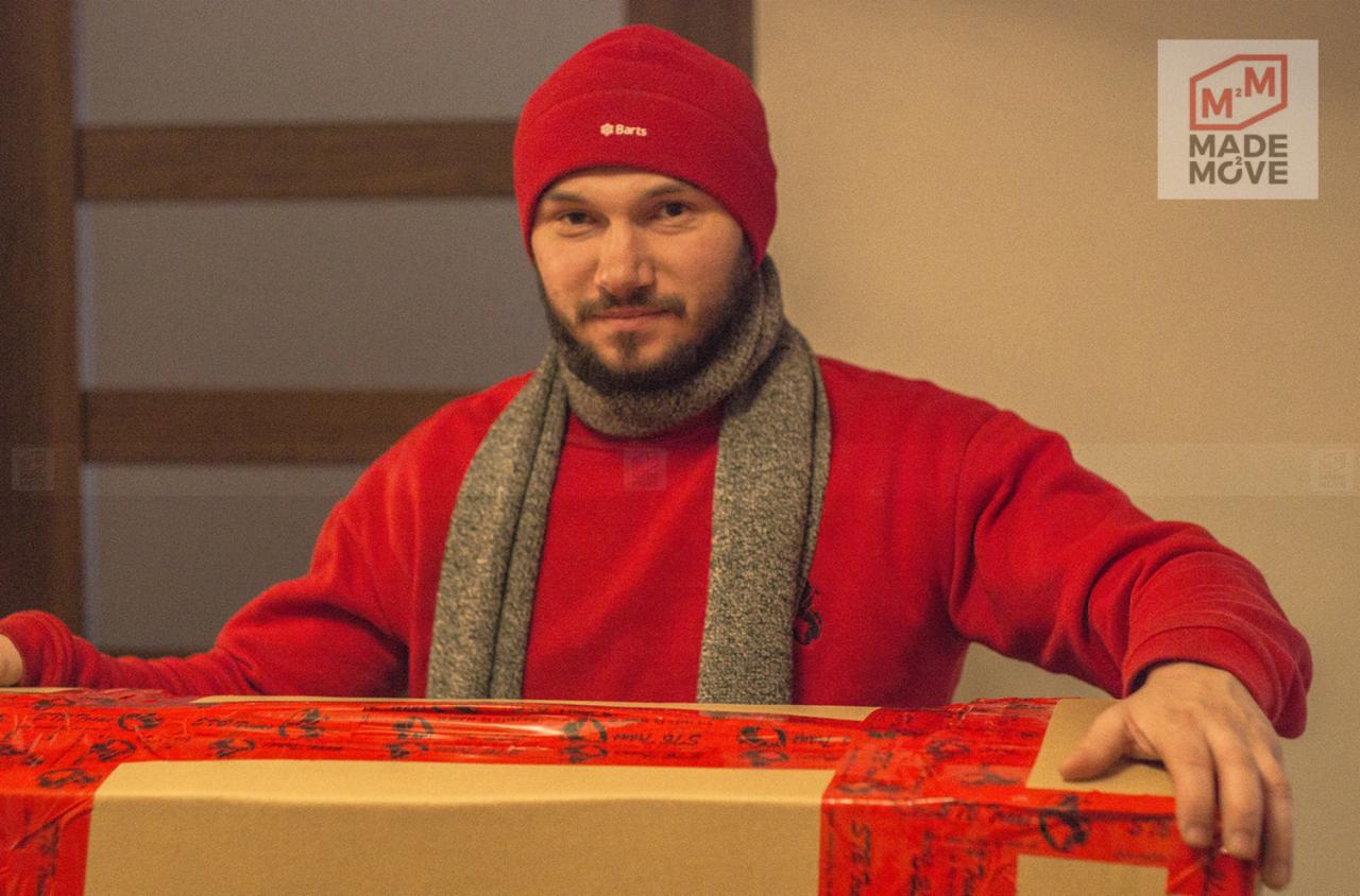 Mężczyzna w czerwonej czapce i swetrze, z szarym szalikiem, opiera ręce na kartonowym pudle zabezpieczonym czerwoną taśmą, widoczne logo firmy w prawym górnym rogu.