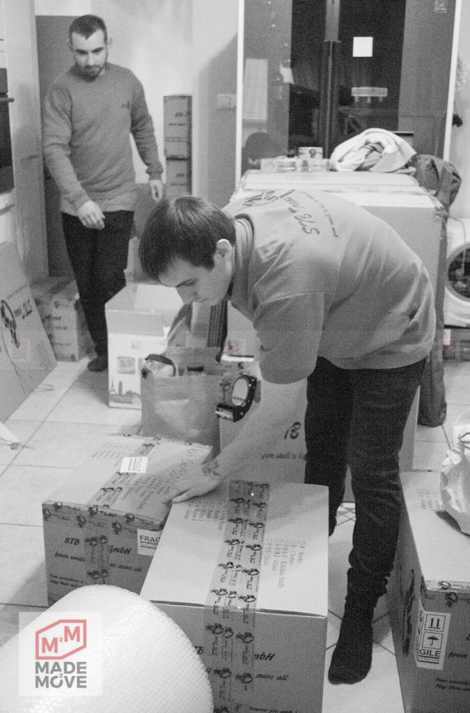 Dwóch mężczyzn w trakcie pakowania kartonów z taśmą klejącą podczas przeprowadzki, widoczne logo firmy na dole po lewej stronie