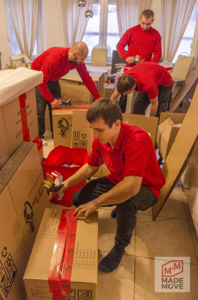 Ekipa w czerwonych koszulkach zakleja taśmą kartony z oznaczeniami pomieszczeń podczas pakowania do transportu, widoczne logo firmy.