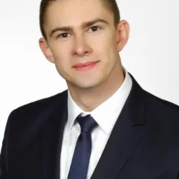 Portret młodego mężczyzny w ciemnym garniturze i białej koszuli z ciemnym krawatem, na jasnym tle.