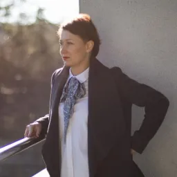 Kobieta w płaszczu i białej koszuli z ozdobnym krawatem, opierająca się o metalową barierkę na tle betonowej ściany w słoneczny dzień.