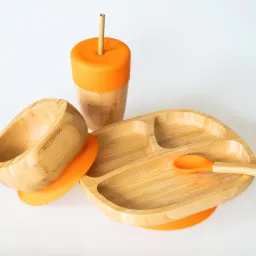 Zestaw naczyń bambusowych dla dzieci: talerzyk trójdzielny, miseczka, kubek z pomarańczową pokrywką i słomką oraz łyżeczka z pomarańczową końcówką, na białym tle.