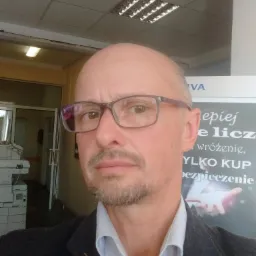 Portret mężczyzny w okularach, z lekkim zarostem, w marynarce i koszuli, na tle biura z kserokopiarką i plakatem reklamowym dotyczącym usług finansowych.