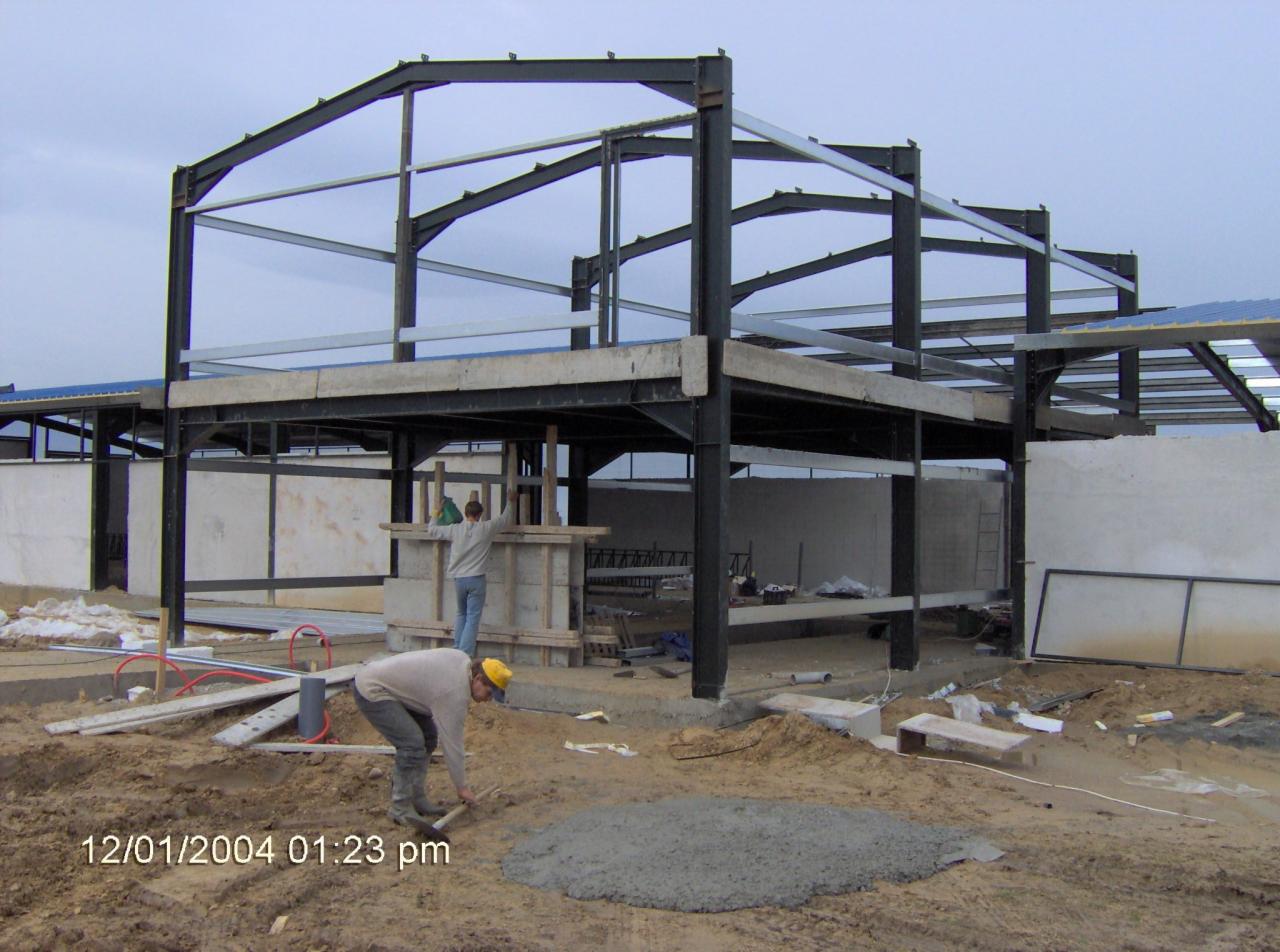 Montaż stalowej konstrukcji hali: pracownik w żółtym kasku rozprowadza beton, w tle widoczny szkielet budynku z dwoma robotnikami przy szalunku.