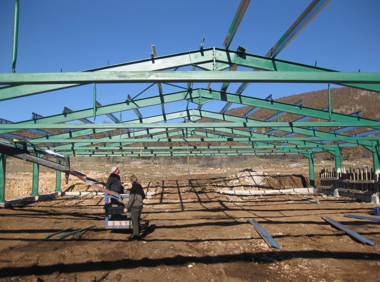 Montaż zielonej konstrukcji stalowej hali na tle wzgórza, dwóch pracowników na podnośniku koszowym, widoczne fundamenty i przygotowany teren.