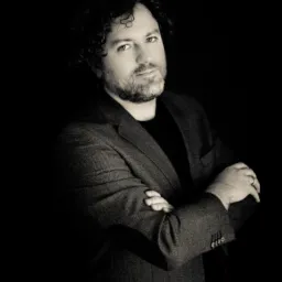 Portret mężczyzny z brodą, w ciemnej marynarce i czarnej koszulce, z założonymi rękami, na ciemnym tle.