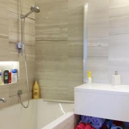 Kompleksowe-Remonty.pl - Nowoczesna łazienka z wanną obudowaną płytkami imitującymi drewno, szklanym parawanem nawannowym, białą szafką z ręcznikami i drewnianą podłogą.