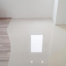 Kompleksowe-Remonty.pl - Białe płytki podłogowe z połyskiem, ułożone w jasnym pomieszczeniu z oknem, widoczny fragment drewnianej podłogi na granicy z płytkami.