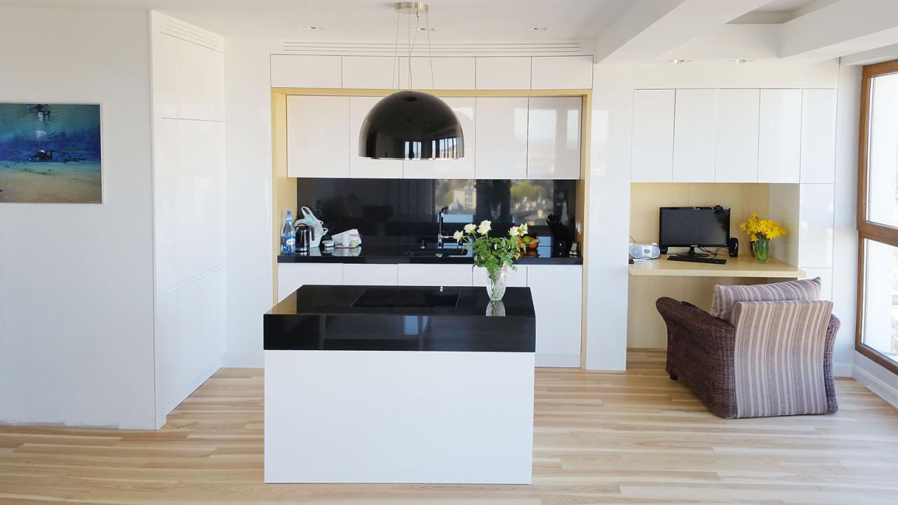 Nowoczesna kuchnia z wyspą o czarnym blacie, białymi szafkami i czarnym panelem nad blatem, uzupełniona o kącik biurowy z komputerem i wiklinowym fotelem.