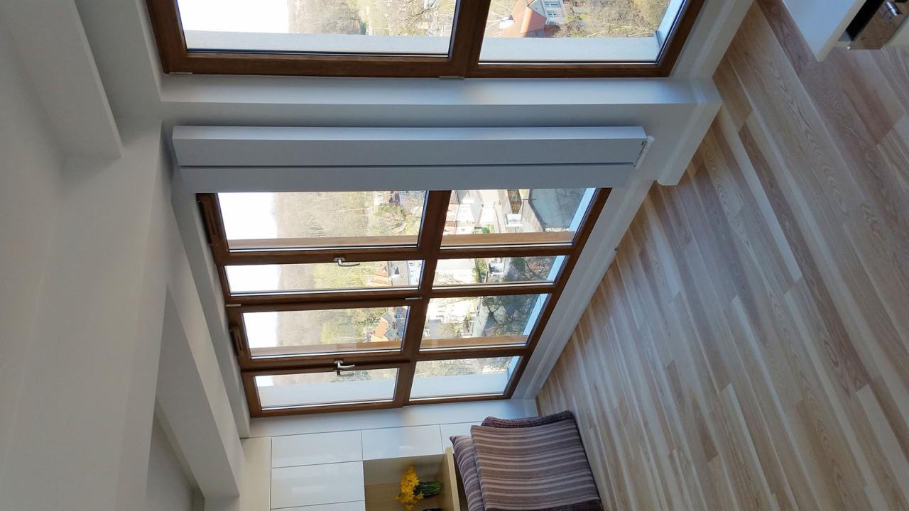 Jasne wnętrze z drewnianą podłogą i dużym oknem dachowym z brązowymi ramami, przez które widać zabudowę miejską. Widoczny fragment białej zabudowy meblowej i dekoracyjne poduszki.