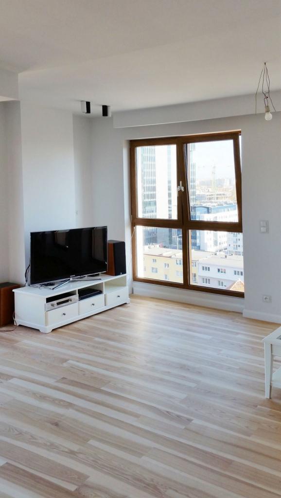 Jasny salon z parkietem w jodełkę, białymi ścianami, dużym oknem z drewnianą ramą i widokiem na miasto. Telewizor na białej szafce RTV.