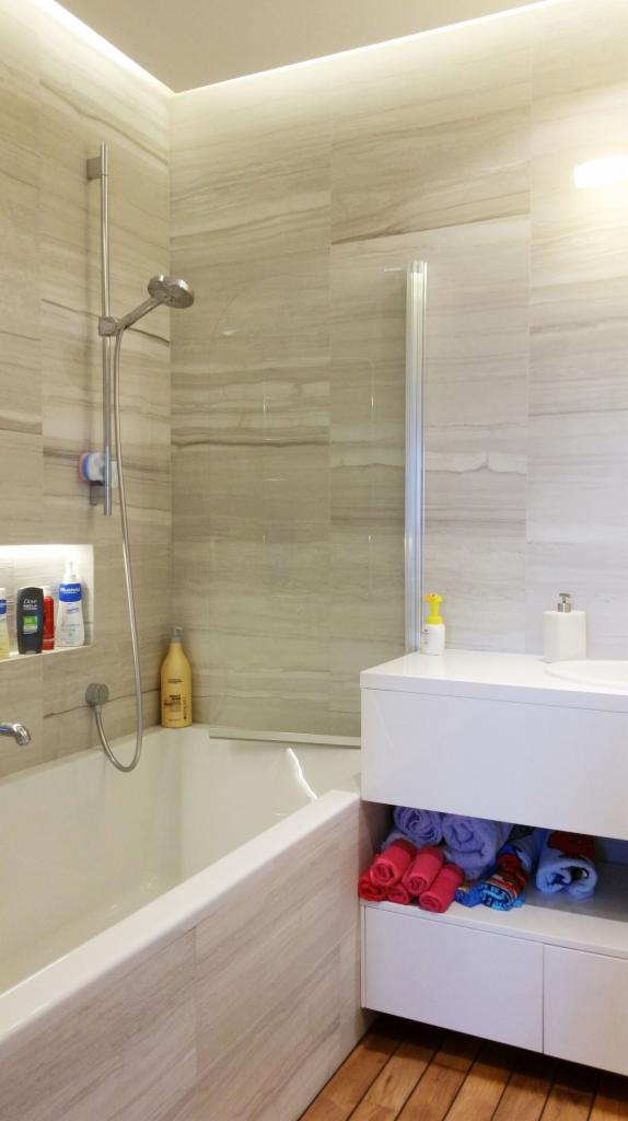 Nowoczesna łazienka z wanną obudowaną płytkami imitującymi drewno, szklanym parawanem nawannowym, białą szafką z ręcznikami i drewnianą podłogą.