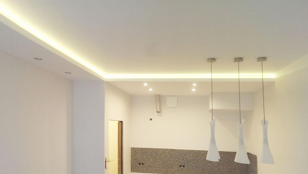 Nowoczesna kuchnia z podświetlanym sufitem LED, minimalistycznymi lampami wiszącymi i mozaiką na ścianie, widok na otwarte przejście do innego pomieszczenia.