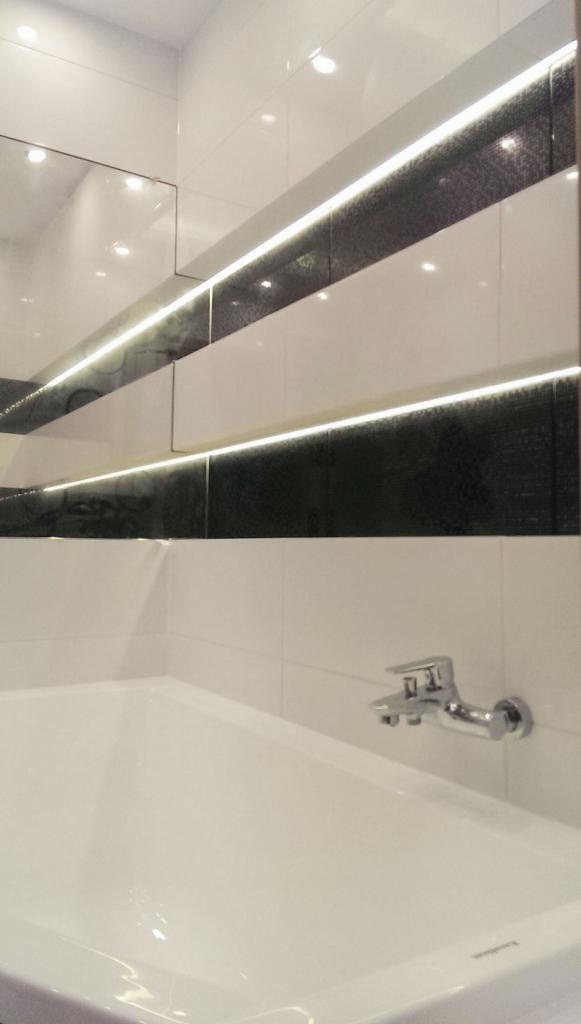 Biała wanna z chromowaną baterią na tle białych płytek z czarnym pasem oświetlonym ledami, nowoczesna aranżacja łazienki.