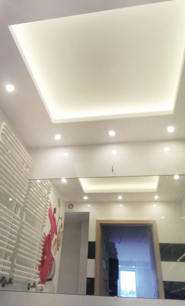 Nowoczesna łazienka z podwieszanym sufitem z oświetleniem LED, grzejnikiem drabinkowym i lustrem odbijającym drzwi do innego pomieszczenia, z dekoracyjną naklejką na ścianie przedstawiającą kwiaty.