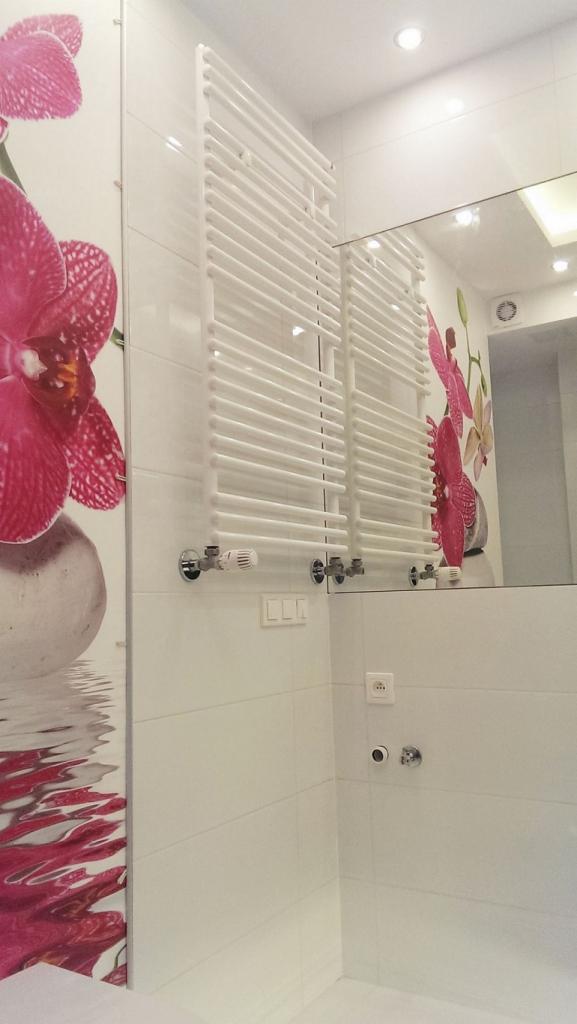 Biała łazienka z grzejnikiem drabinkowym, płytkami i dużym lustrem, dekorowana motywem kwiatowym na ścianie.