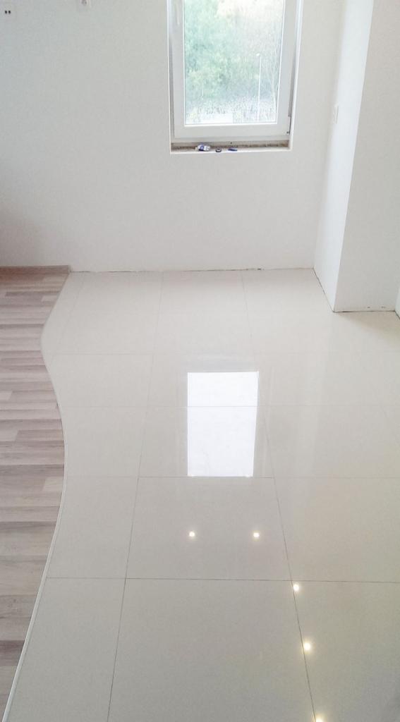 Białe płytki podłogowe z połyskiem, ułożone w jasnym pomieszczeniu z oknem, widoczny fragment drewnianej podłogi na granicy z płytkami.