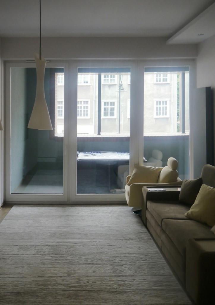 Nowoczesny salon z dużym oknem balkonowym, szary dywan z fakturą drewna, beżowy fotel obrotowy i brązowa sofa z poduszkami w odcieniach żółci i brązu, widok na balkon z łóżkiem.