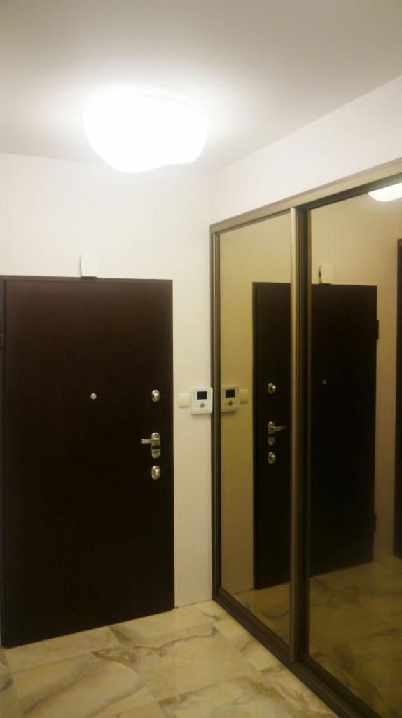 Ciemne drzwi wejściowe z wizjerem i srebrną klamką obok przesuwnych drzwi szafy wnękowej z lustrem, na marmurowej podłodze, oświetlone plafonem.