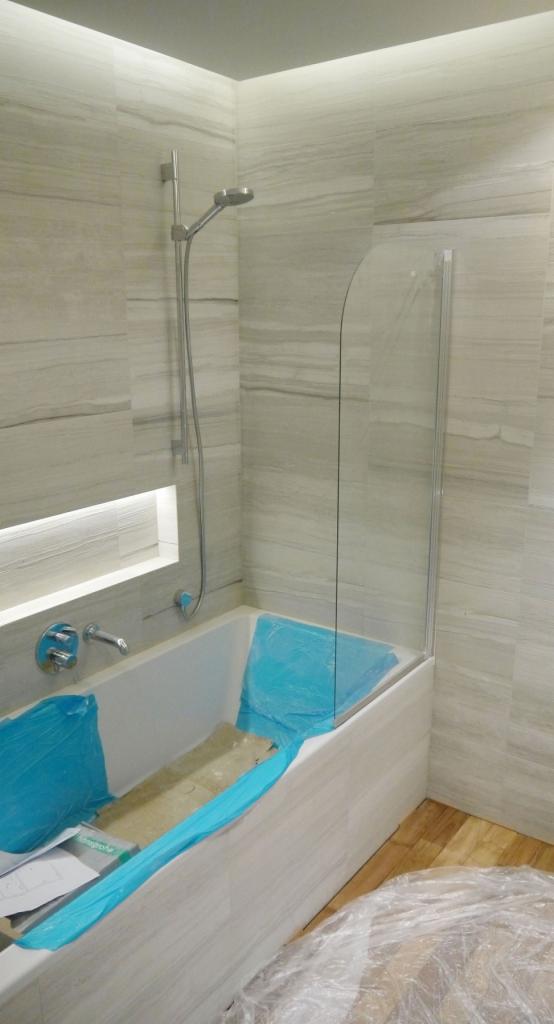 Półokrągła szklana ścianka prysznicowa zamontowana na wannie, zabezpieczona folią, ściany wyłożone płytkami imitującymi drewno, drewnopodobna podłoga częściowo zakryta folią ochronną.